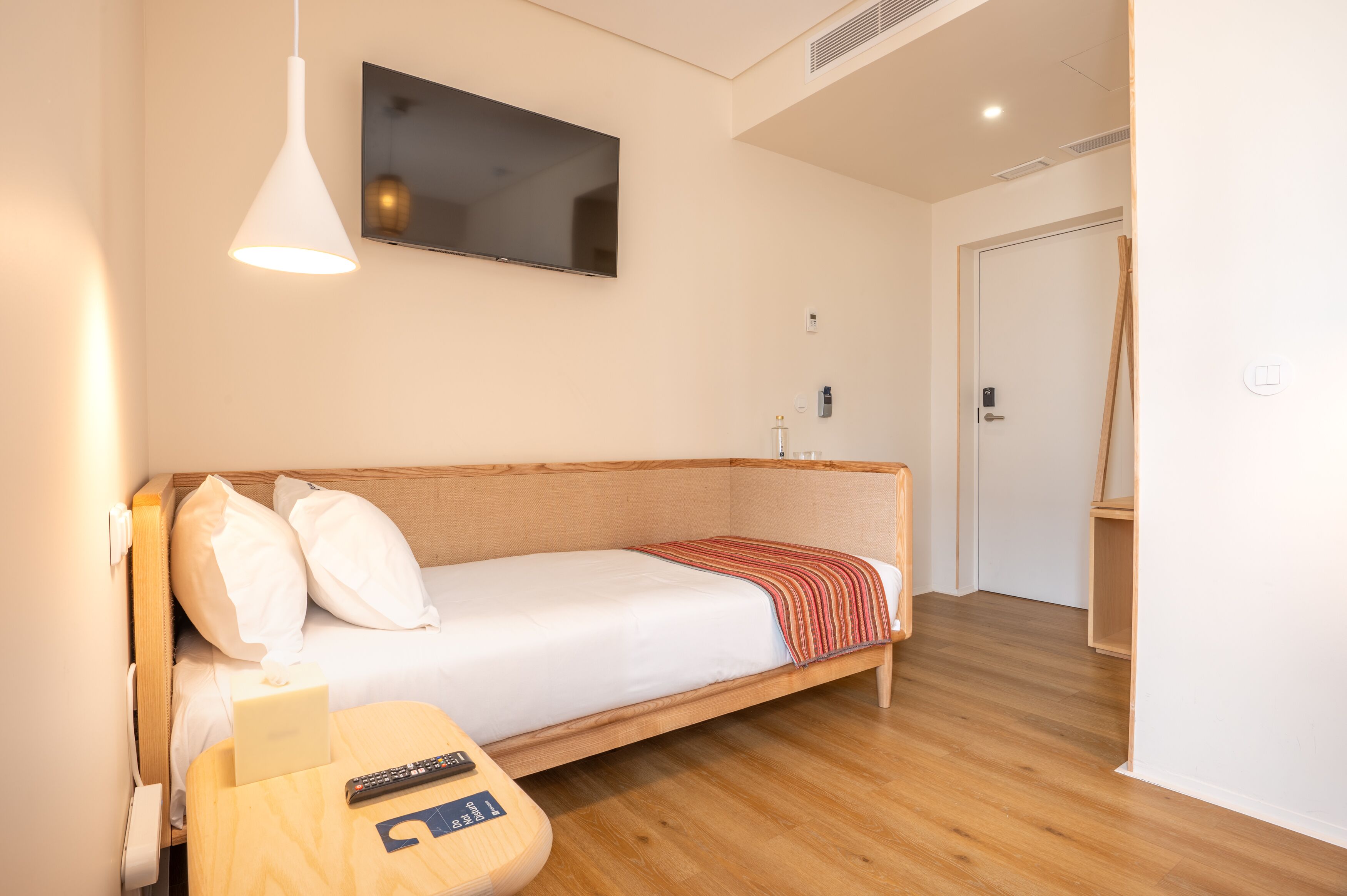 A single bed with a wooden headboard, a bedside table with a remote control, and a mounted TV.