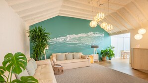 Un salón amplio con techo alto, un gran mural de un pueblo costero, un sofá color beige y varias plantas en macetas.