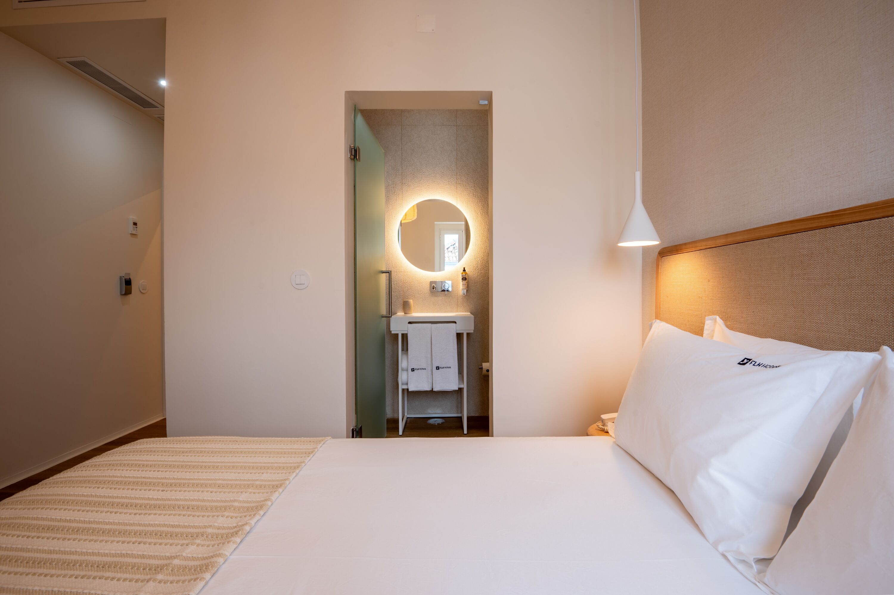 A hotel room with a bed, a bedside table, a nightstand, a wall-mounted round mirror, a small white cabinet, and a hanging light fixture.