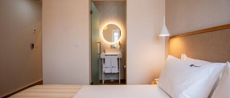 A hotel room with a bed, a bedside table, a nightstand, a wall-mounted round mirror, a small white cabinet, and a hanging light fixture.