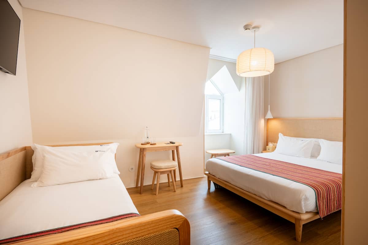 A hotel room with two beds, a small table, a chair, and a window with a view.