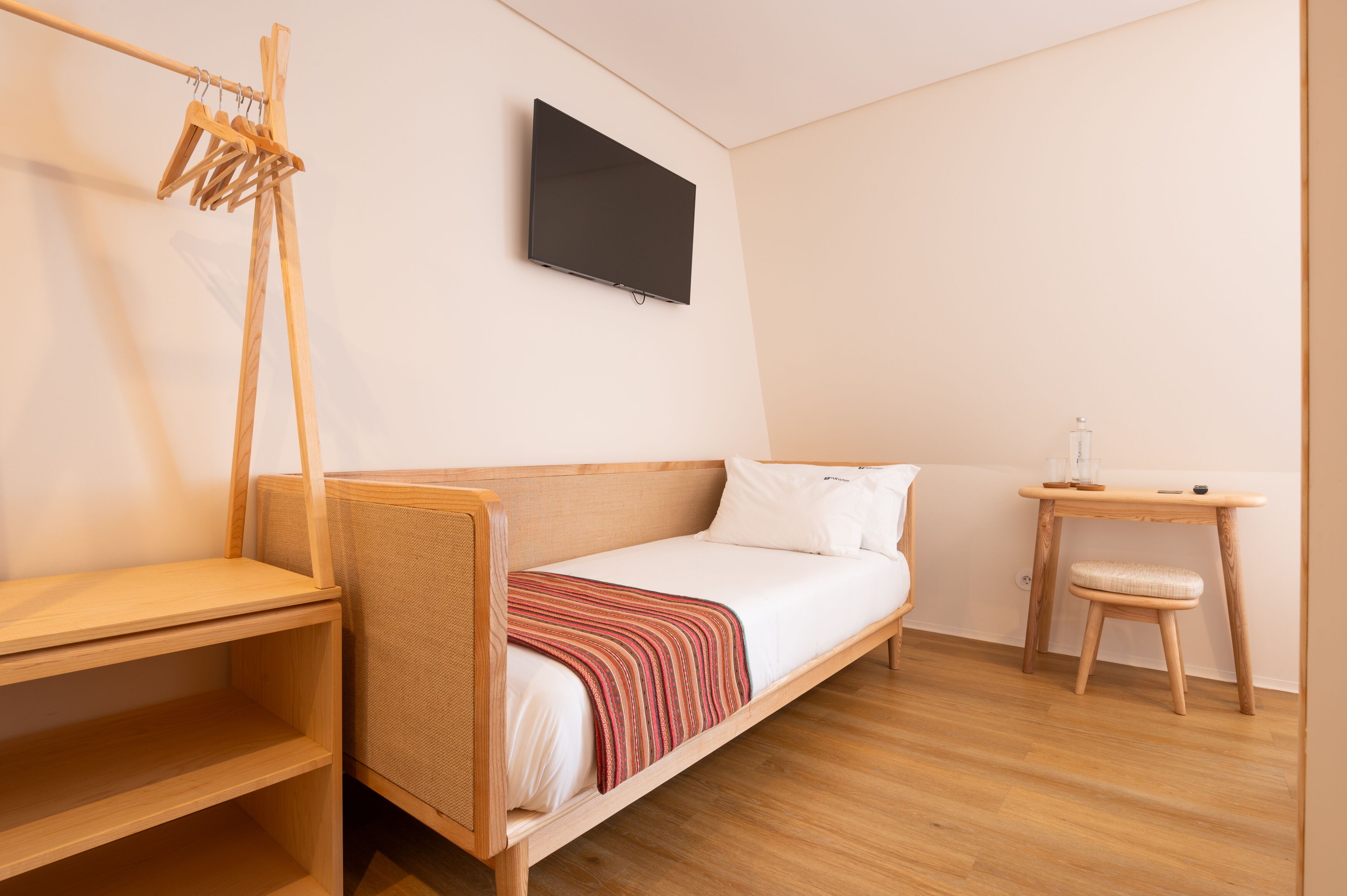 Une petite chambre comprenant un lit simple, une table de chevet en bois, un petit tabouret et un téléviseur fixé au mur.