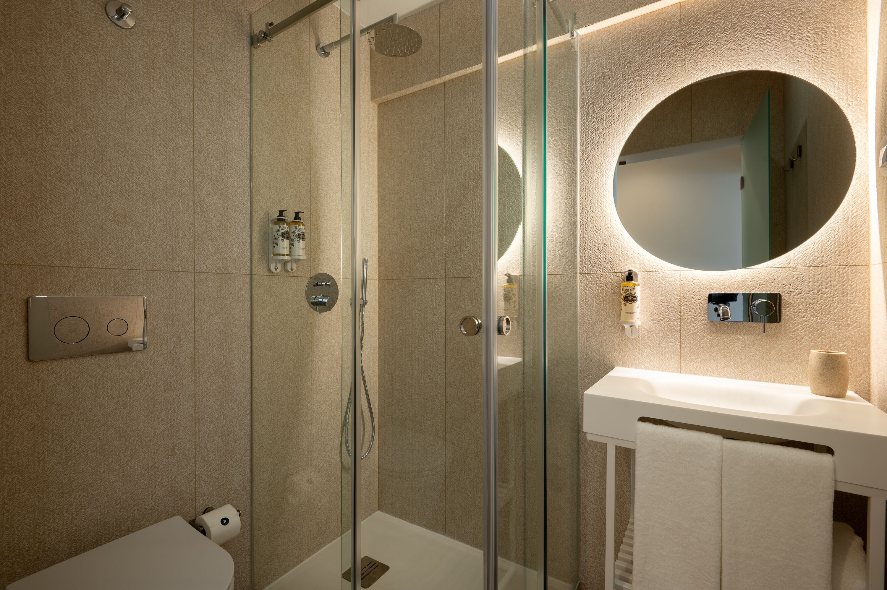 Une salle de bain moderne avec une douche à parois de verre, un miroir rond et un lavabo blanc.