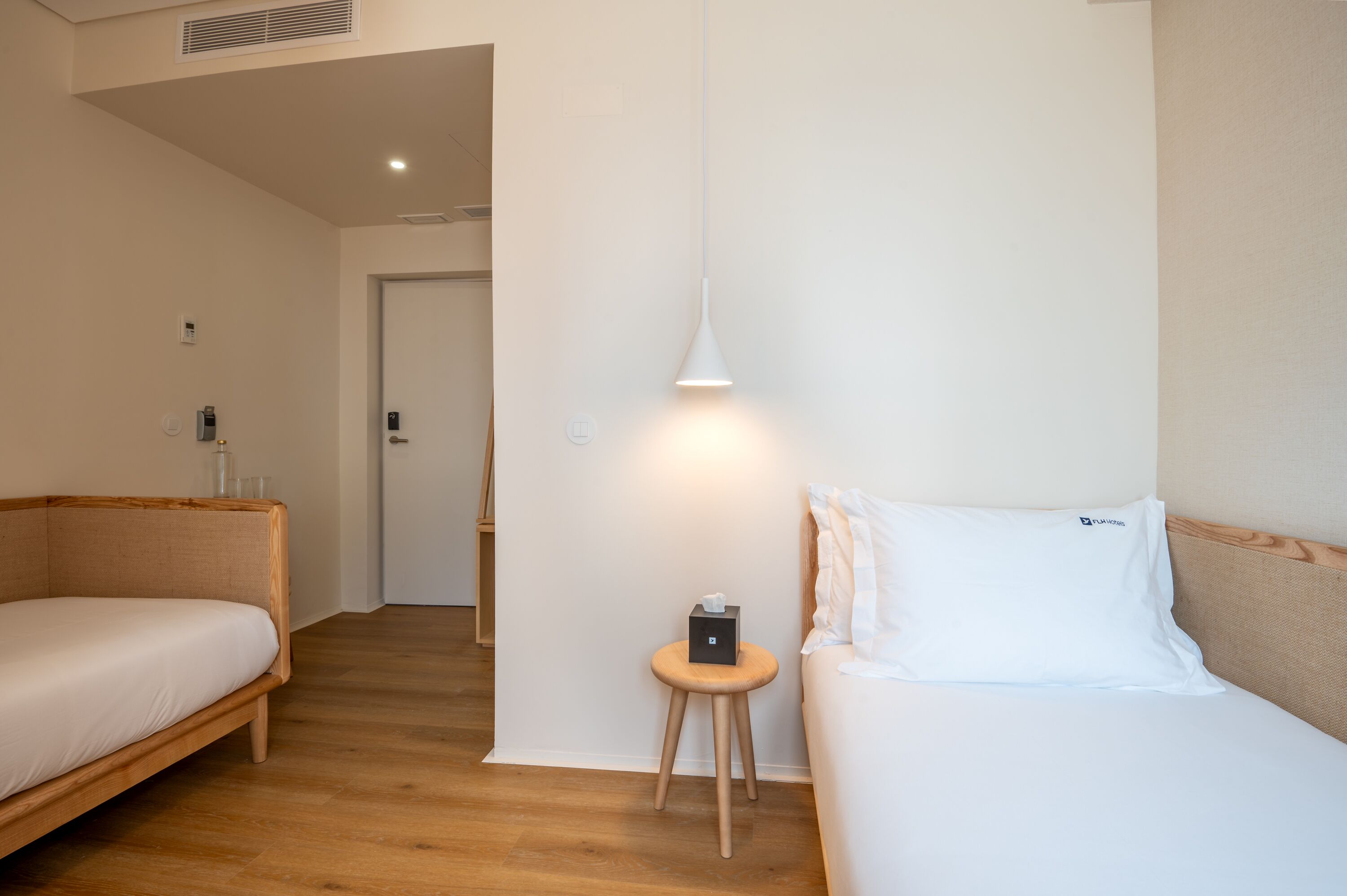 A hotel room with a wooden floor, a bed with white bedding, a bedside table with a lamp, and a door leading to another room.