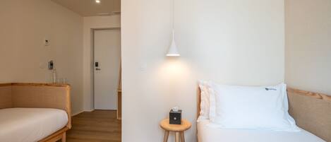 A hotel room with a wooden floor, a bed with white bedding, a bedside table with a lamp, and a door leading to another room.