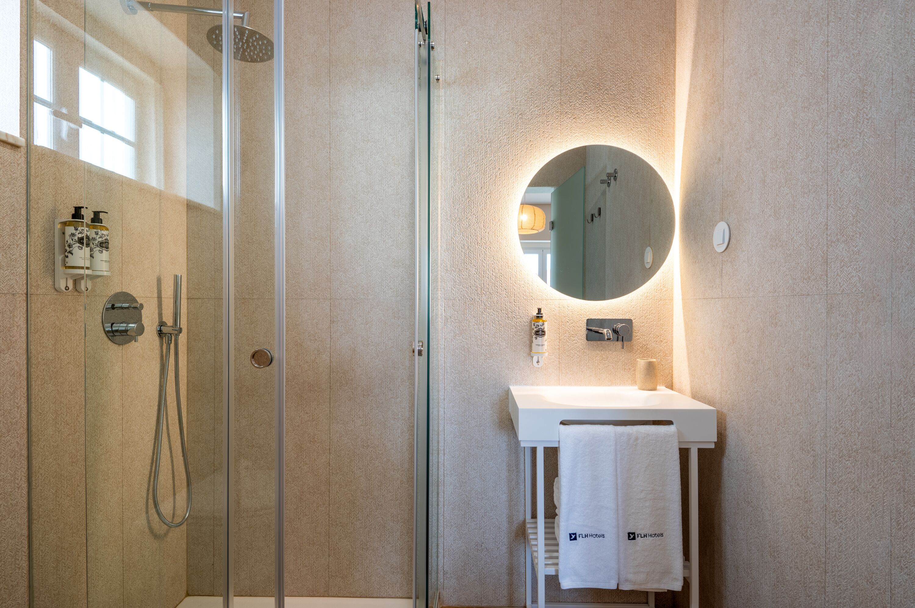 A modern bathroom with a glass-enclosed shower, a round mirror, and a white sink with a towel hanging on a rack.