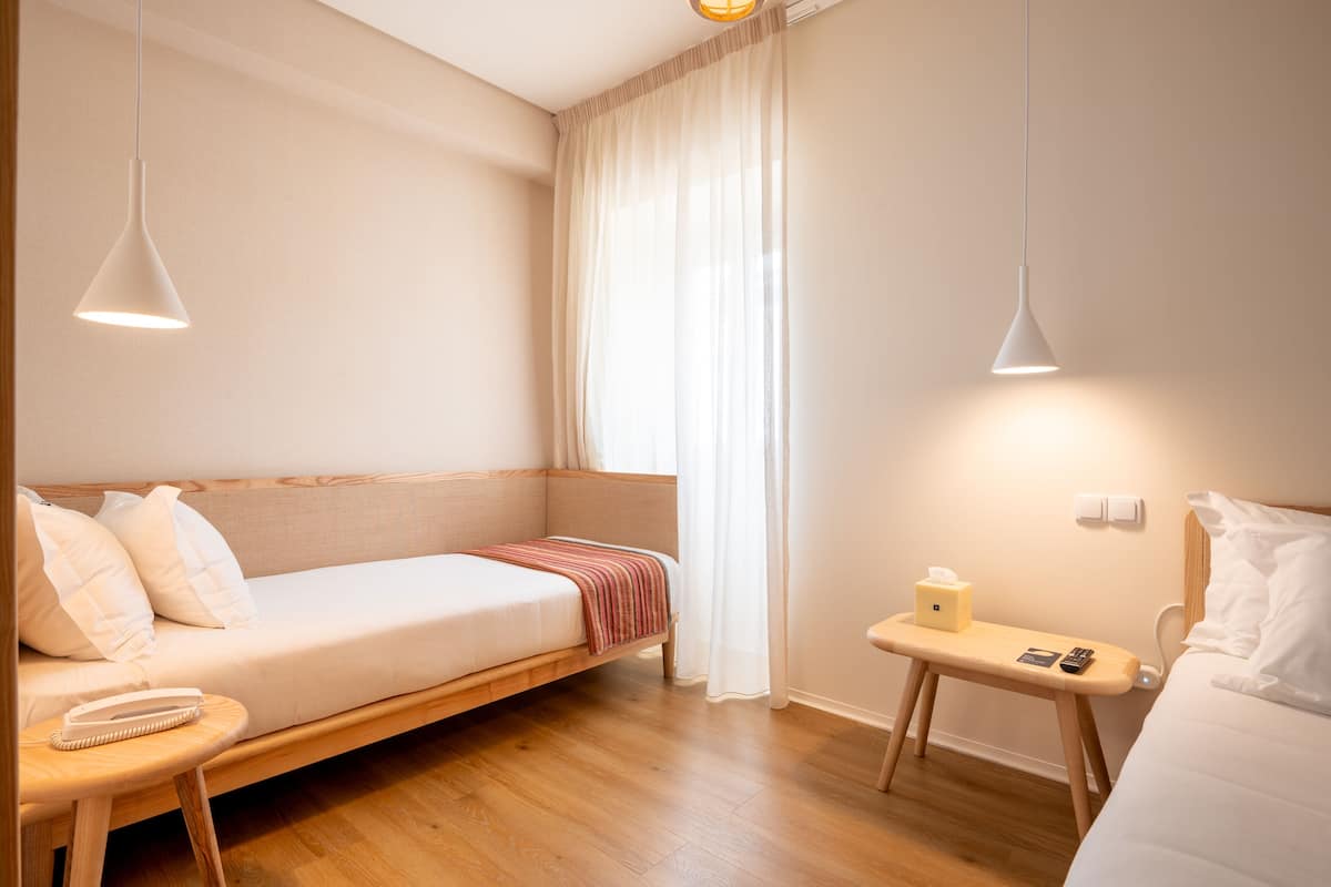 A modern hotel room with a wooden floor, a sofa bed, a bedside table with a lamp, and a small bedside table with a tissue box and remote control.
