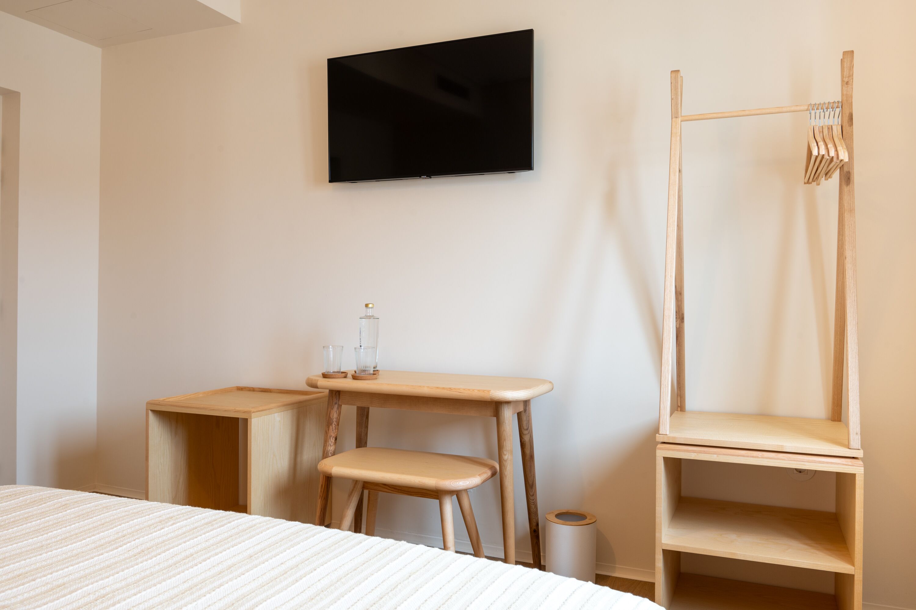 Une chambre d’hôtel minimaliste comprenant un lit, une table de chevet en bois, un téléviseur fixé au mur et une armoire en bois.