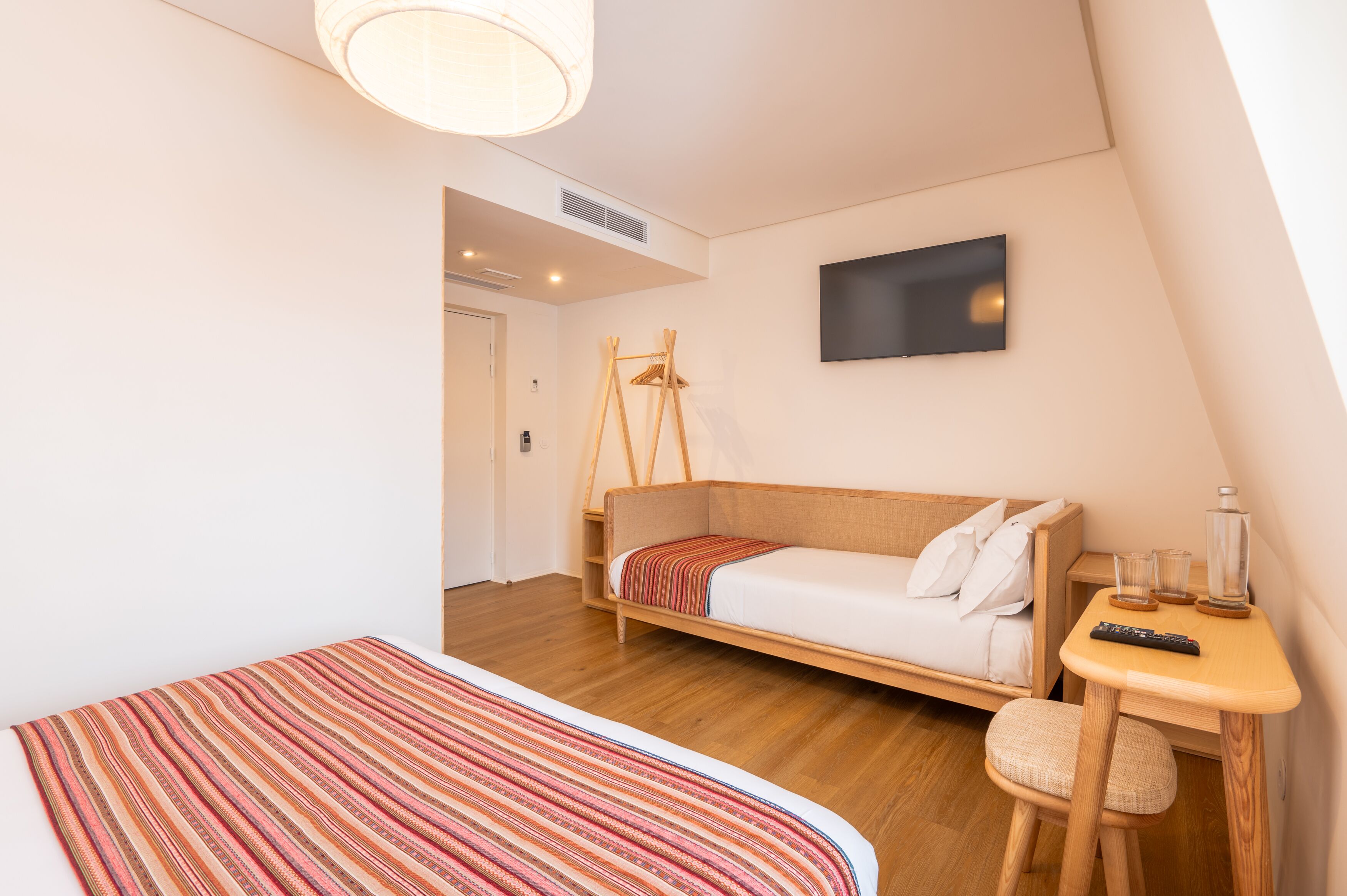 A modern hotel room with a wooden floor, a bed with a striped blanket, a small table with a remote control, and a TV on the wall.
