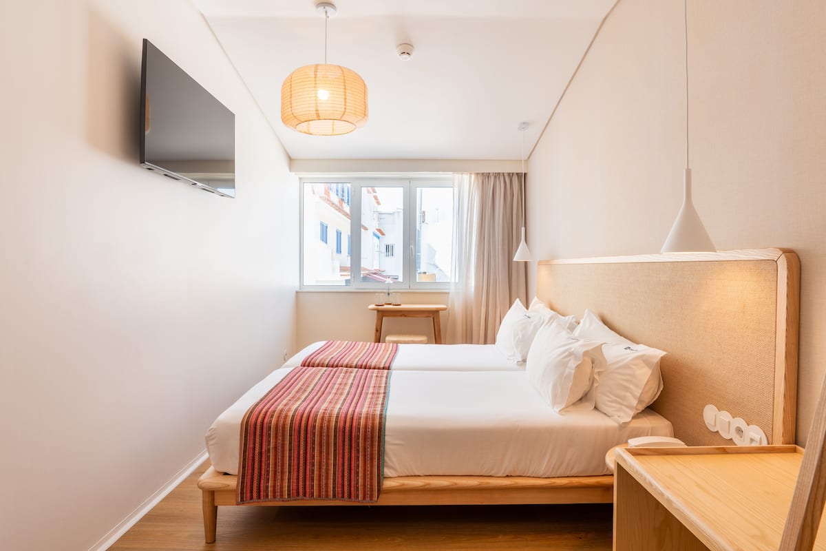 A hotel room with a bed, a bedside table, a TV mounted on the wall, and a window with curtains.