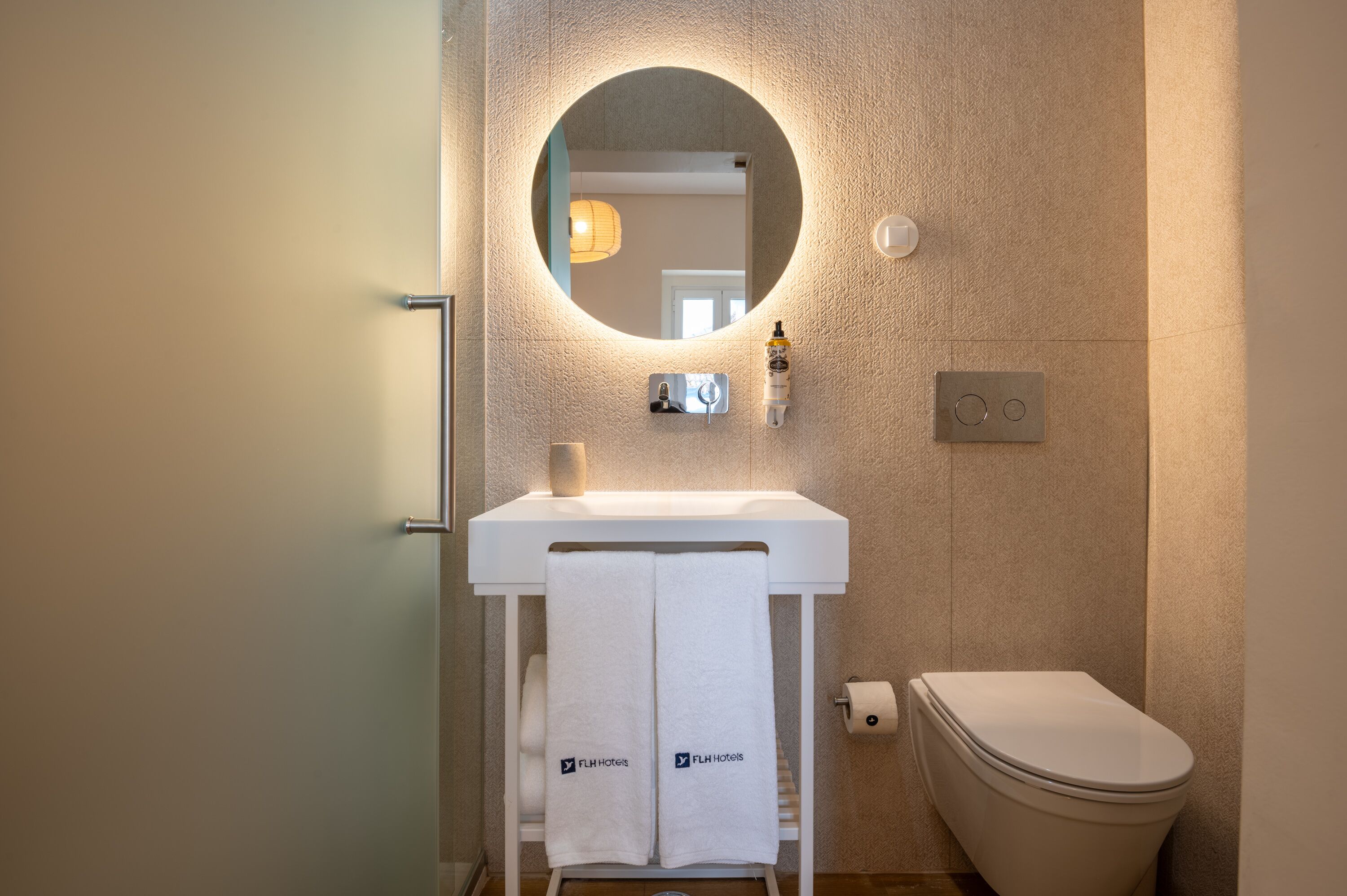 Een moderne badkamer met een ronde spiegel, een wit wastafelmeubel en een toilet. Handdoeken hangen aan een rek en er staat een fles handzeep op de wastafel.