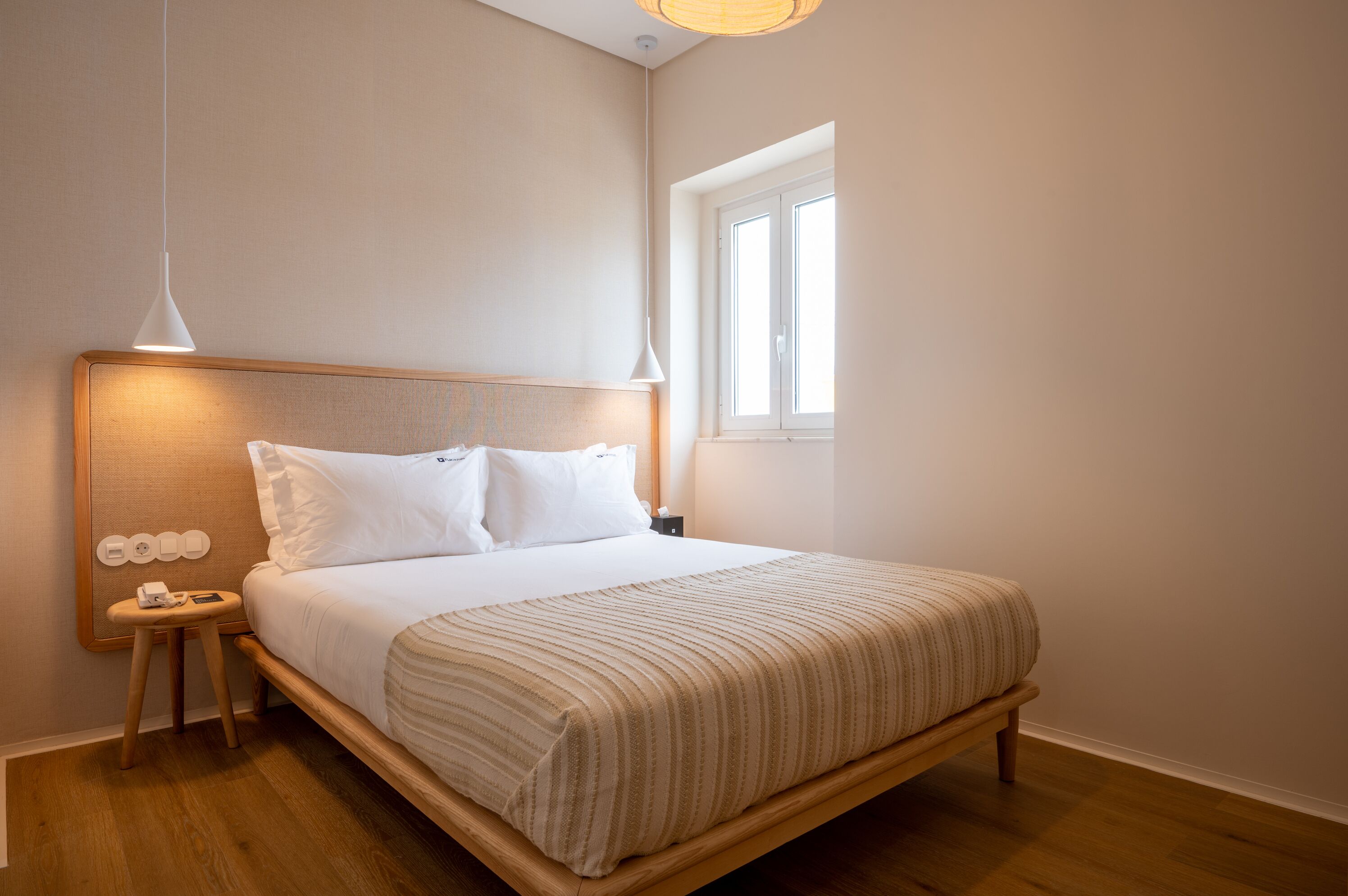 A neatly made bed with a wooden headboard, a bedside table with a lamp, and a window providing natural light.