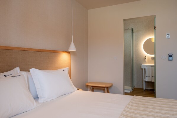 Habitación de hotel con cama, cabecero de madera, mesita de noche, taburete pequeño y baño visible a través de una puerta de cristal.