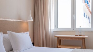 Una habitación de hotel con una cama, una mesita de noche con lámpara, una ventana con cortinas y vistas a un edificio.