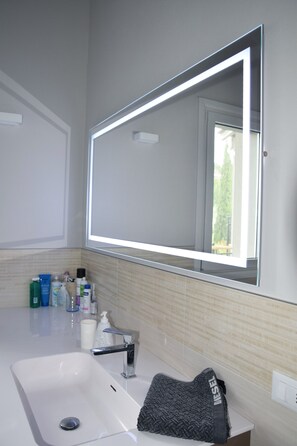 A bathroom with a white sink, a mirror with a light strip, and toiletries on the counter. - La Bella Vita, a new three-room apartment with a pool, just a few minutes from Lake Garda. (Boschetti)