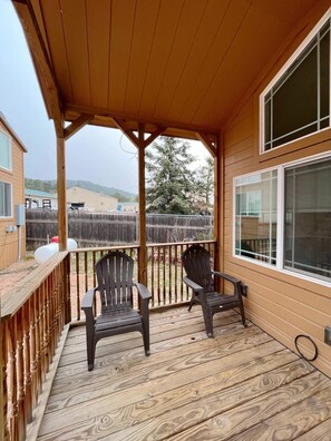 A covered deck with two Adirondack chairs, a wooden railing, and a view of a fenced yard with a tree and buildings in the background. - Tiny home Within Campground (Woodland Park)