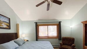 A bedroom with a bed, a chair, a ceiling fan, a window with curtains, a nightstand, and a lamp.