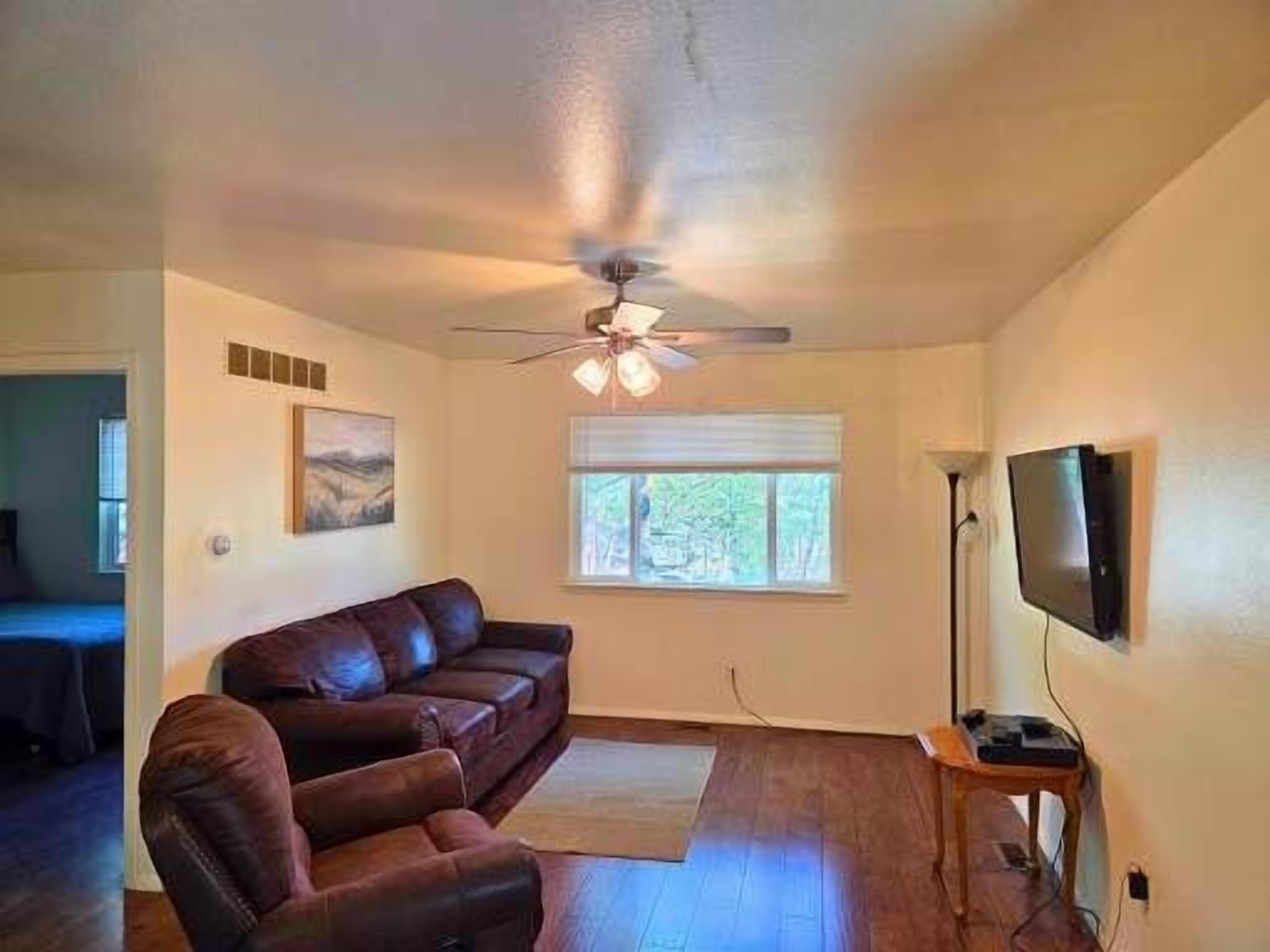 Una sala de estar con un sofá de cuero marrón, un ventilador de techo, un televisor en la pared y una ventana con persianas.