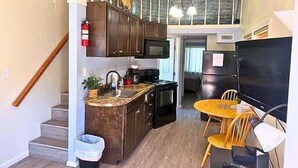 A compact kitchen with a microwave, stove, and sink, a dining area with a table and chairs, and a staircase leading to an upper level.