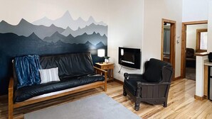 A cozy room with a wooden ceiling, a ceiling fan, a black sofa, a grey rug, a wooden coffee table, a TV, and a mountain mural on the wall. - Cabin Suite Bed & Breakfast (Woodland Park)