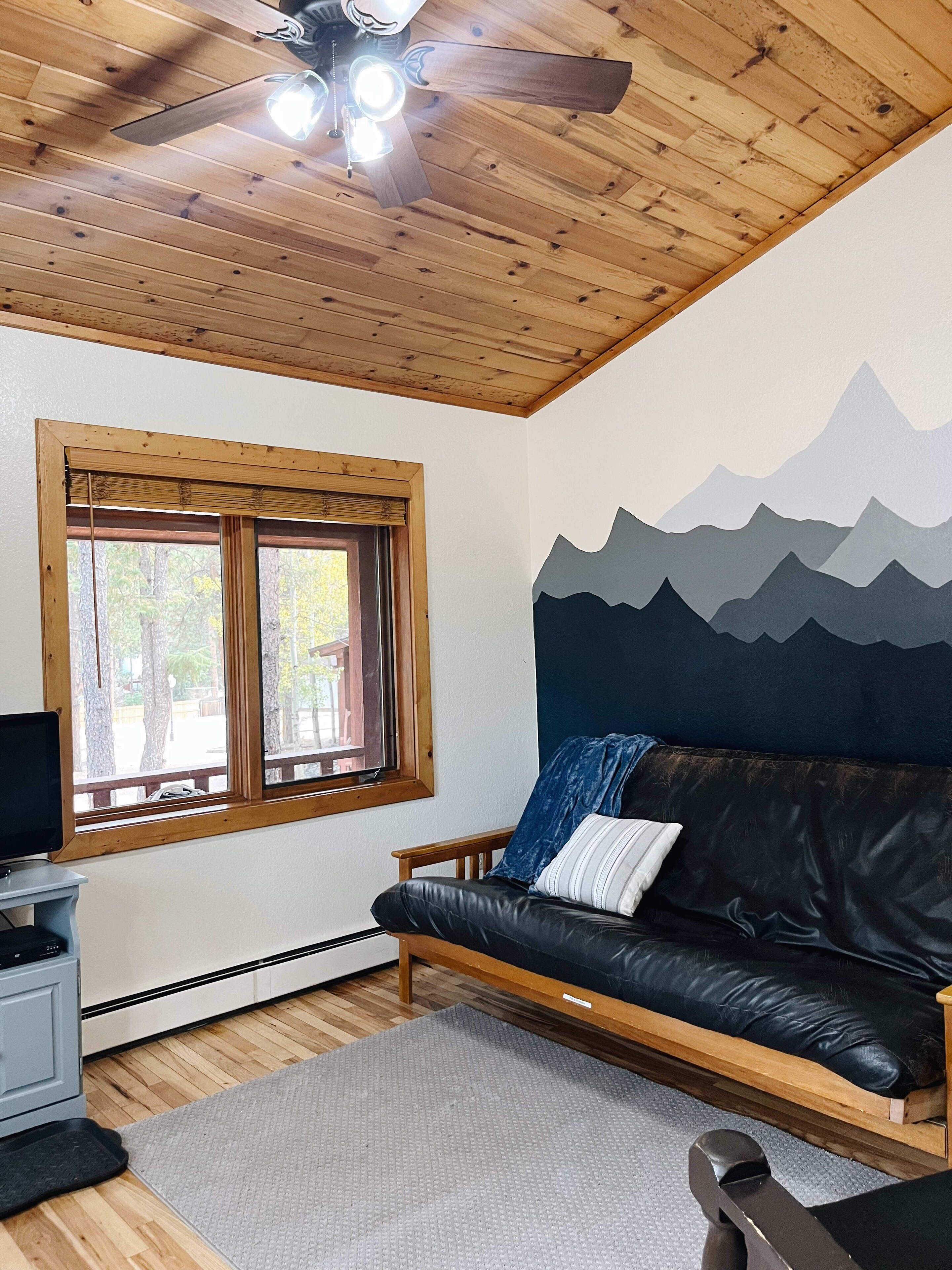 A room with a wooden ceiling, a black leather sofa, a window with a view of trees, and a wall mural of mountains.