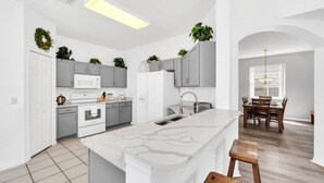 Une cuisine moderne avec des meubles gris, un îlot central blanc et un coin repas doté de chaises en bois et d’un lustre.
