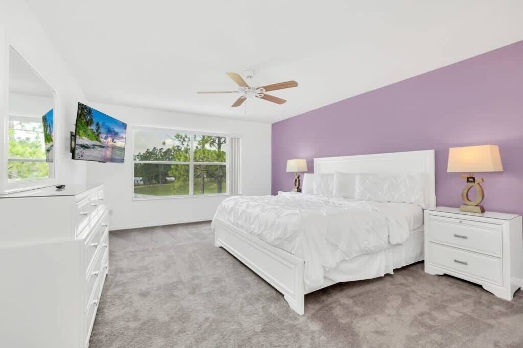 A bedroom with a bed, two bedside tables, a ceiling fan, a TV on the wall, and a window with a view of greenery.
