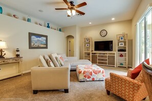 A living room with a sofa, ottoman, and armchair, a ceiling fan, a television, and built-in shelving.