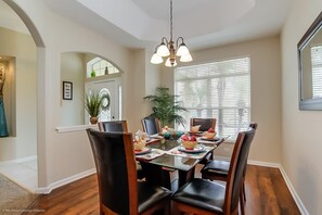 A dining room with a wooden table set for six, surrounded by chairs. There is a chandelier above the table, and a large window with blinds to the right.