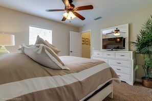 A bedroom with a bed, a ceiling fan, a dresser, a mirror, a lamp, and a potted plant.