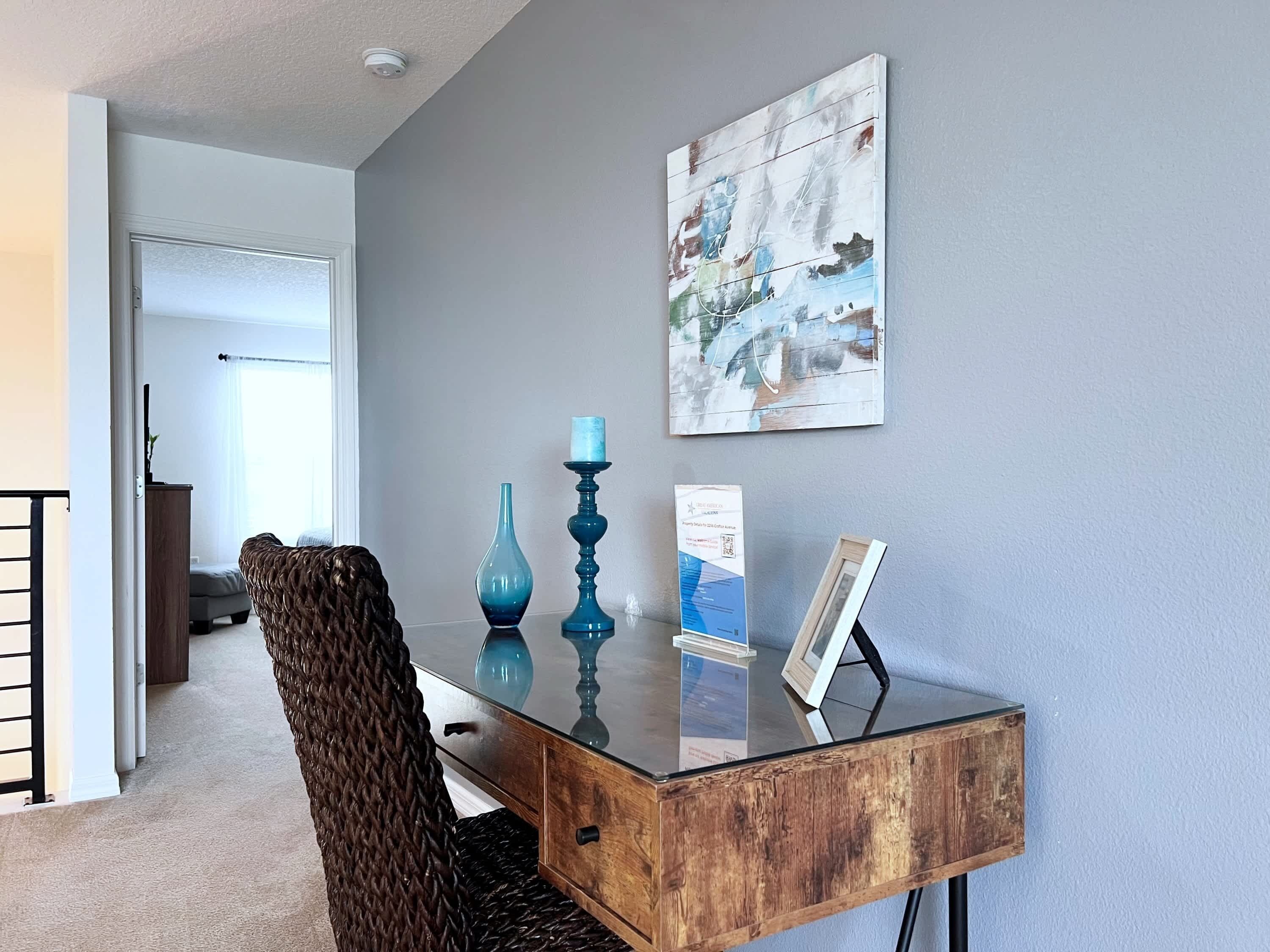 A modern office space with a glass desk, a woven chair, a blue vase, and a framed picture on the wall.