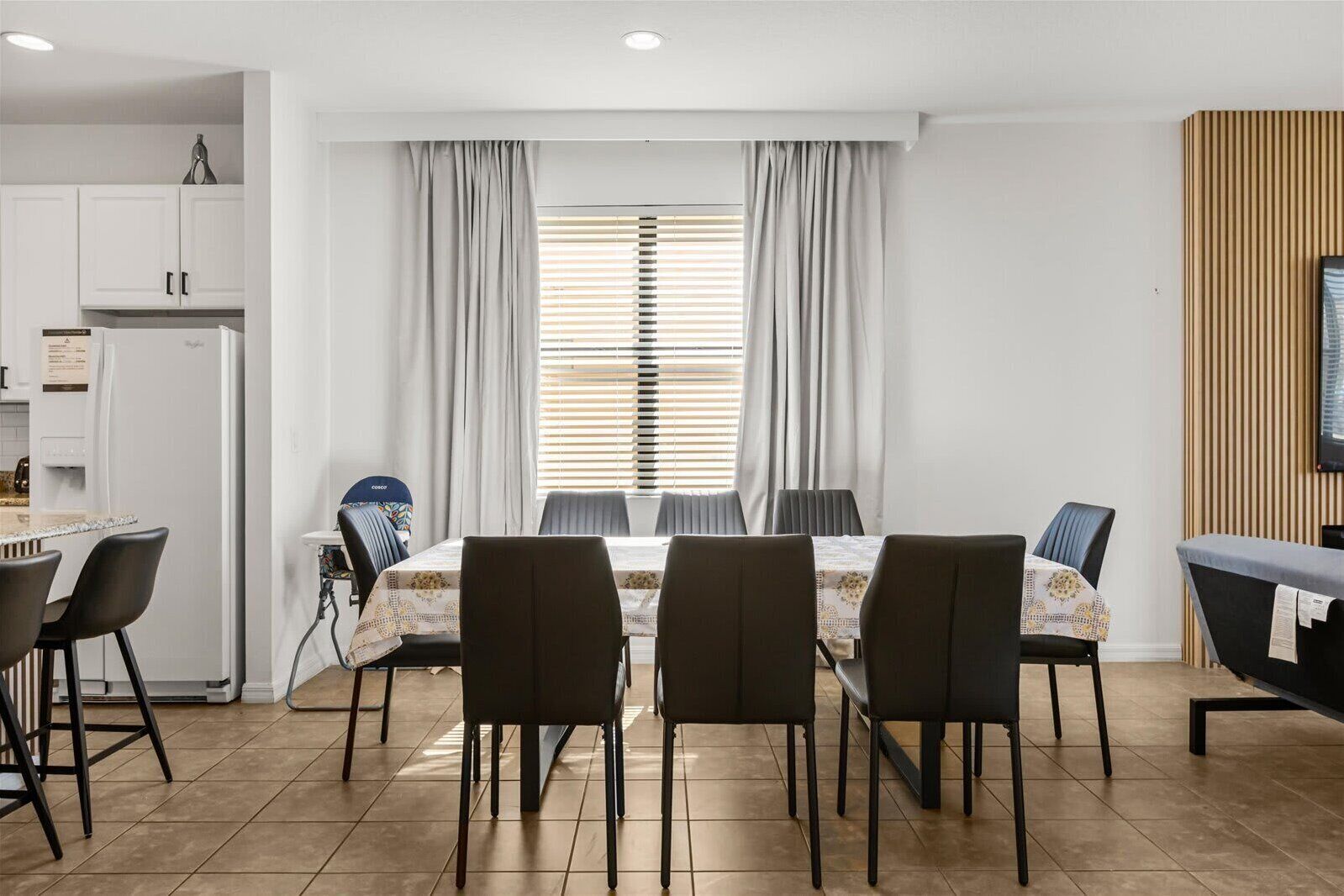 A dining area with a table set for four, a kitchen with white cabinets, and a window with blinds.