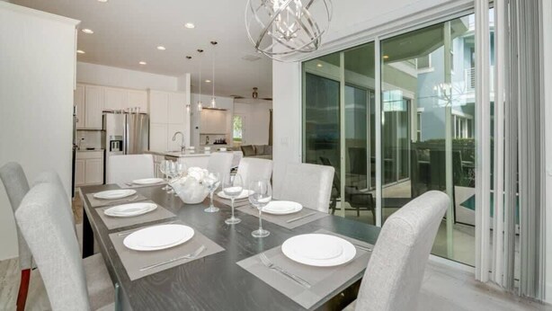A modern dining area with a long table set for six, white chairs, and a contemporary chandelier. - Private Pool | 15 Min to Disney | Gated Resort 1472 (Kissimmee)
