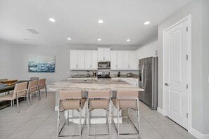 A modern kitchen with white cabinets, a central island with bar stools, a dining area with a table and chairs, and a large refrigerator.