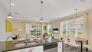 Eine moderne Küche mit Geschirrspüler, einem Essbereich mit weißen Stühlen und einem großen Fenster mit Jalousien.