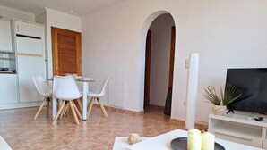 A modern living room with a white sofa, a dining table with chairs, a TV on a stand, and a small plant. - VF012 Apartament LaBella Cumbre del Sol - Benitachell, Costa Blanca (Benitachell)
