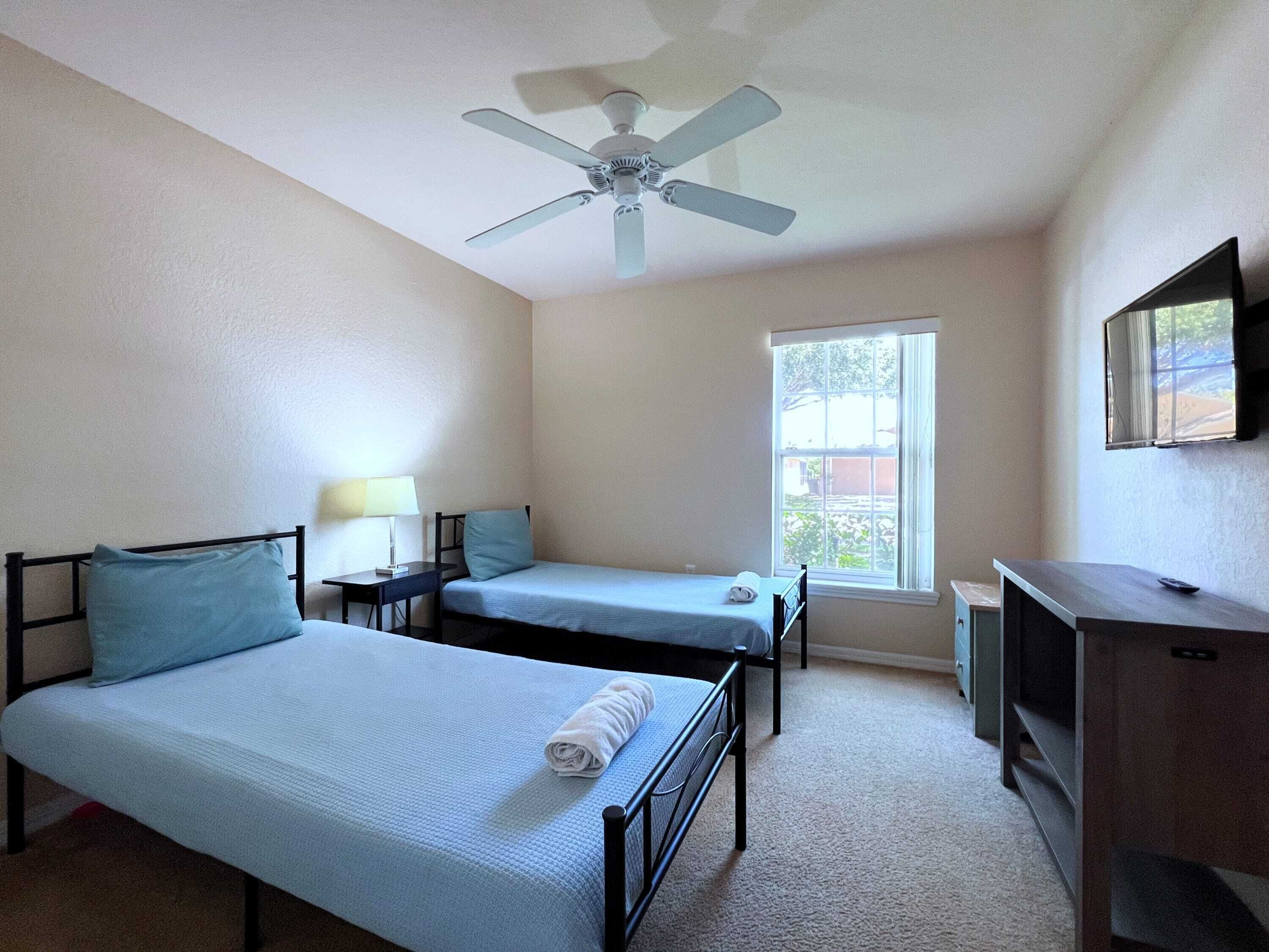 Ein Zimmer mit zwei Einzelbetten, einem Schreibtisch, einem Stuhl, einem Deckenventilator, einem Fenster und einem an der Wand montierten Fernseher.