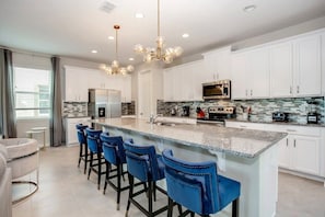 Une cuisine moderne avec des meubles blancs, un grand îlot central doté de tabourets de bar bleus et un lustre.