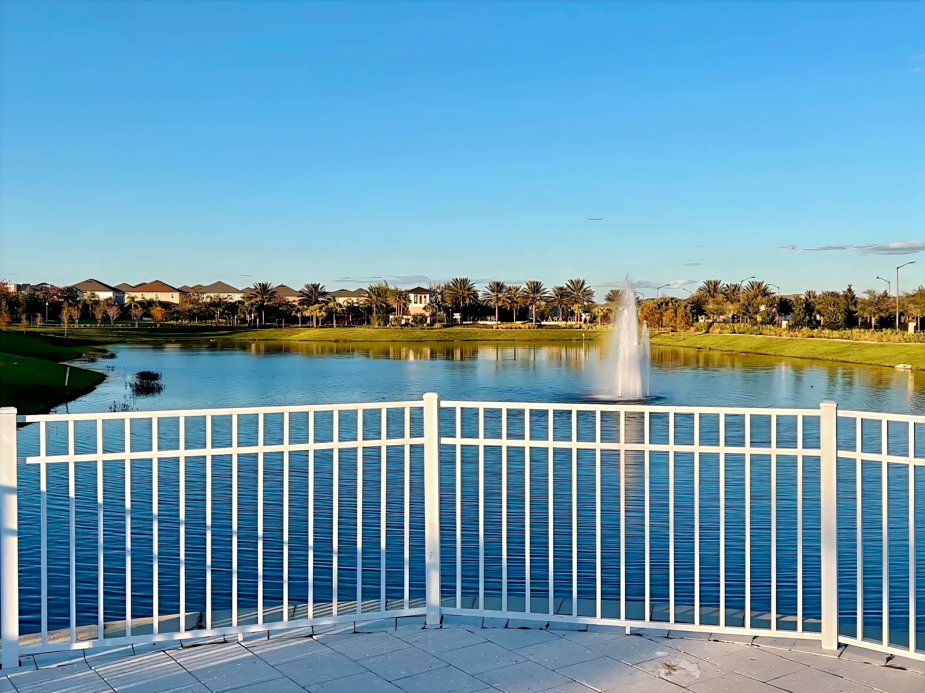 Una vista tranquila de un lago con una fuente, una cerca blanca y un cielo despejado.