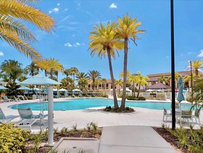 A pool area with lounge chairs and umbrellas, surrounded by palm trees and a clear blue sky.