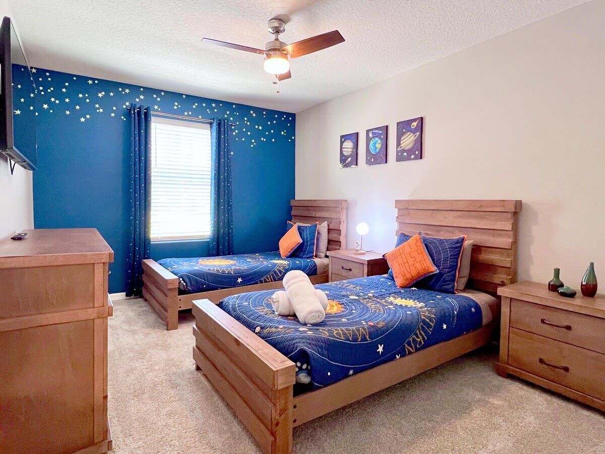 Quarto com duas camas de madeira, um ventilador de teto, cômoda e janela com cortinas.