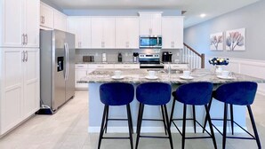 Una cocina moderna con armarios blancos, electrodomésticos de acero inoxidable, encimera de mármol y taburetes de bar azules.
