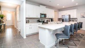 Una cocina moderna con armarios blancos, una isla central con taburetes de bar, electrodomésticos de acero inoxidable y salpicadero de azulejos.