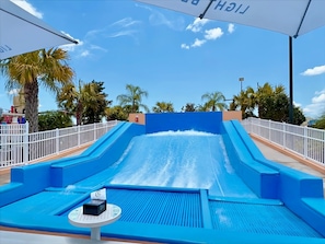 A water slide with a blue pool at the bottom, surrounded by white railings and palm trees.