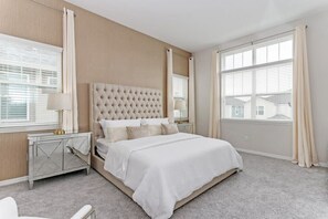 A bedroom with a tufted headboard, a large window with blinds, a bedside table with a lamp, and a carpeted floor. - Harry Potter | Harley Game Loft | Near Disney 1787 (Kissimmee)