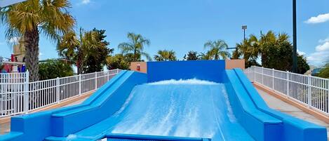 A water slide with a blue pool at the bottom, surrounded by palm trees and a white fence.