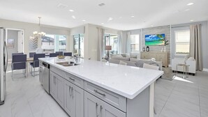 Una cocina moderna con una isla grande, un comedor con sillas y una sala de estar con sofá y televisor.