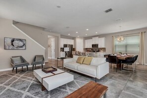 A spacious living room with a white sofa, two armchairs, a coffee table, and a dining area with a table and chairs.