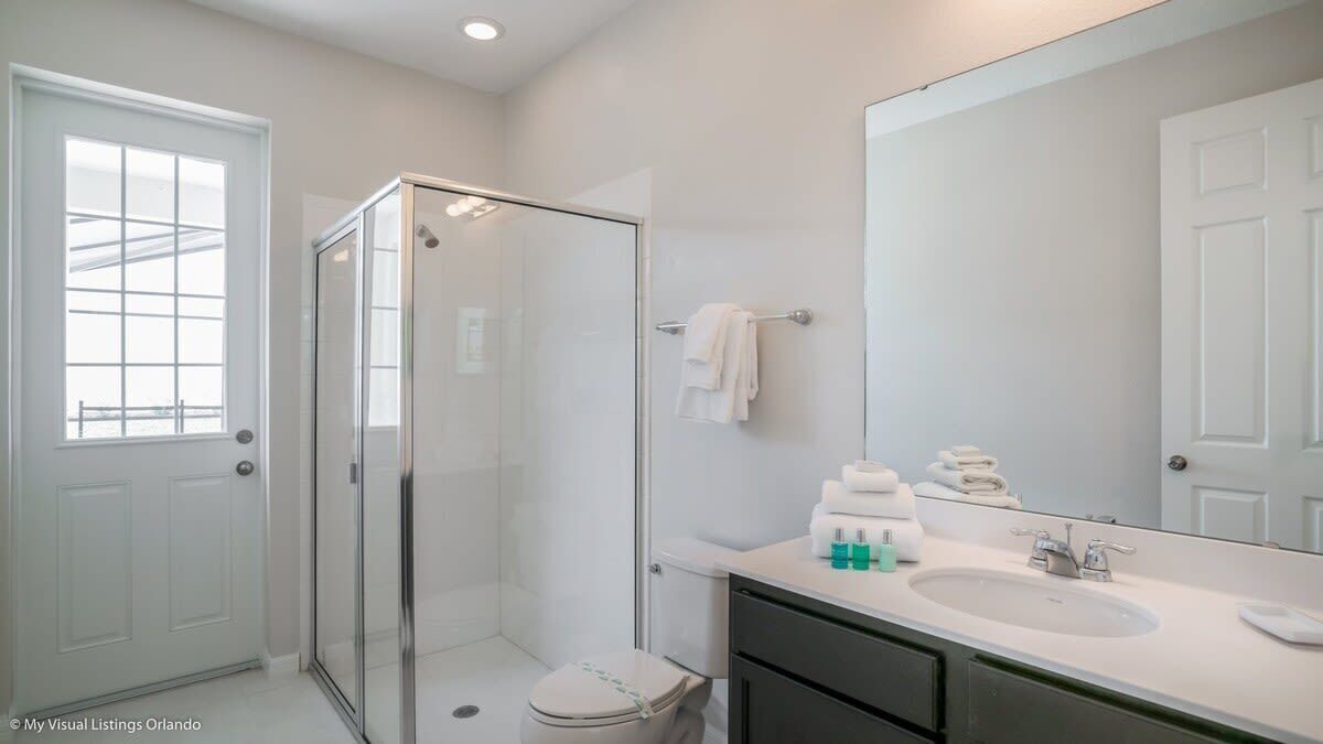 Un baño moderno con cabina de ducha de vidrio, inodoro blanco, lavamanos con artículos de aseo y un espejo grande.