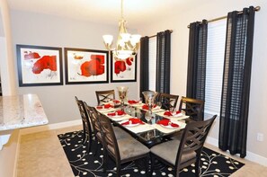 A dining area with a rectangular table set for four, surrounded by chairs. There are framed artworks on the wall above the table, a chandelier hanging from the ceiling, and a window with curtains. - No Rear Neighbors | Resort | Pool | Game Room 4183 (Davenport)